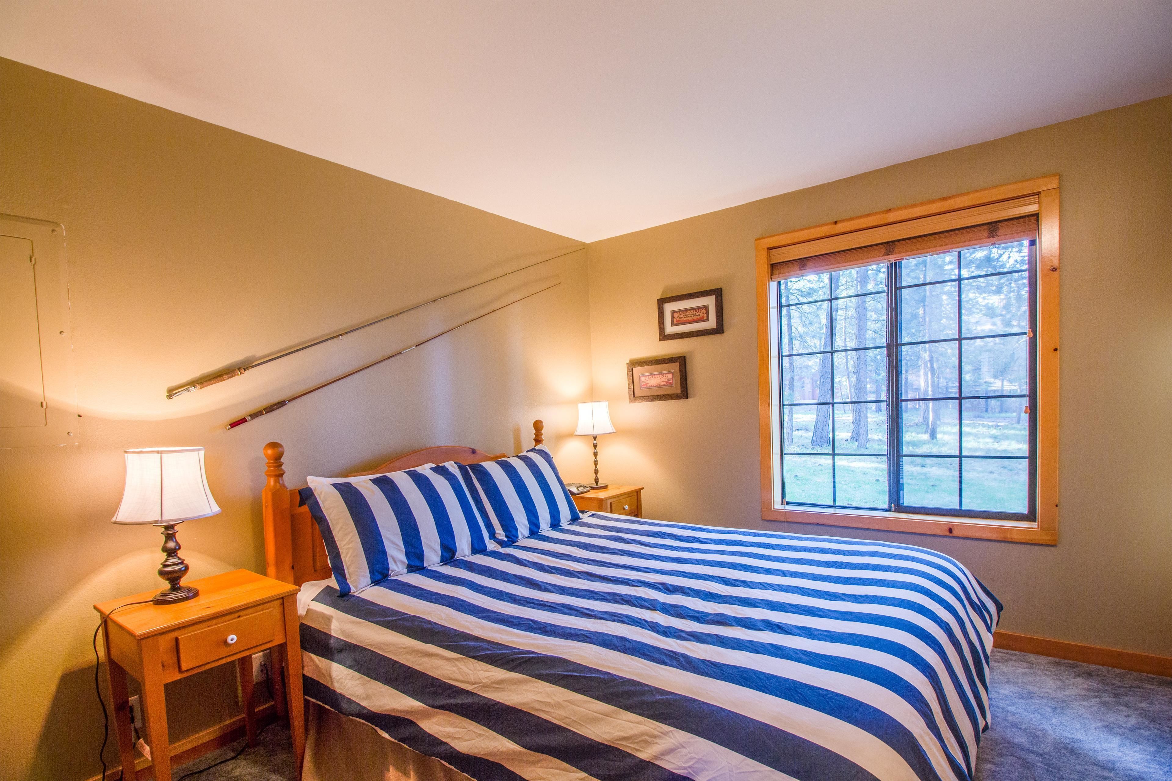 A bedroom with a bed, a nightstand with a lamp, a window with a view of trees, and a framed picture on the wall.
