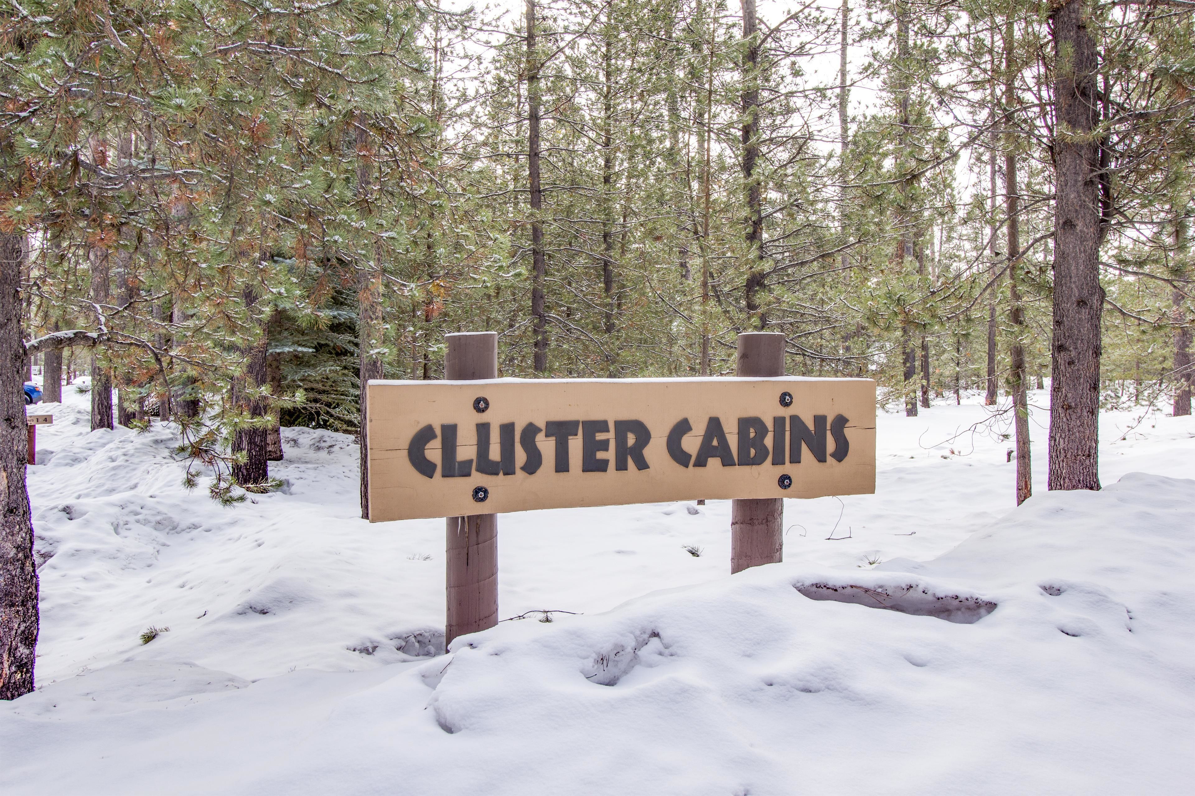 Un panneau en bois portant l’inscription « CLUSTER CABINS », placé dans un décor de forêt enneigée.