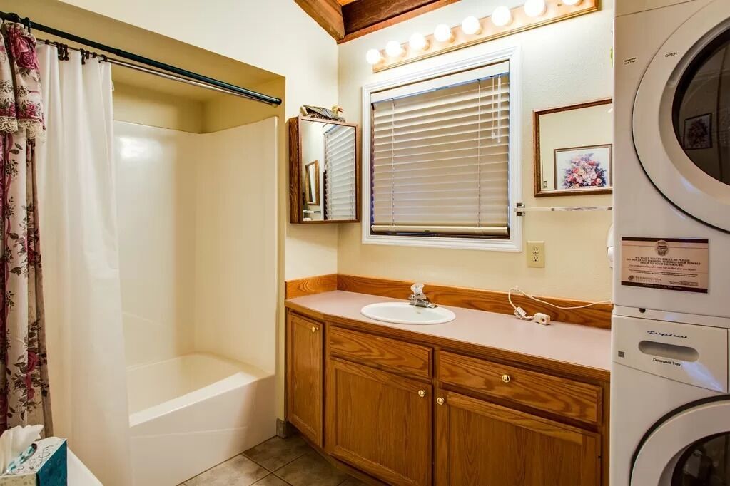 Ein Badezimmer mit einem Holzkosmetikschrank, einer Badewanne, einer Waschmaschine und einem Fenster mit Jalousien.