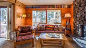 Un interior acogedor de cabaña con revestimiento de madera, una chimenea, una estantería y un amplio ventanal con vistas al exterior.