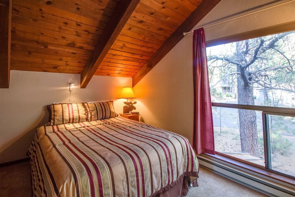 Quarto com teto de madeira, uma janela ampla com vista para árvores, cama com roupa de cama listrada e mesa de cabeceira com abajur.