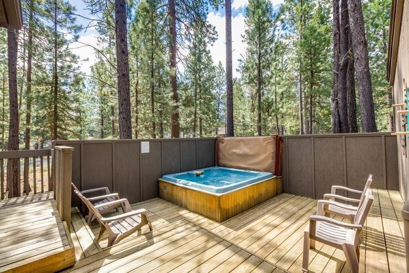 A wooden deck with a hot tub, two chairs, and a view of tall pine trees.
