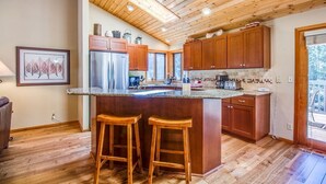 A kitchen with wooden cabinets, a stainless steel refrigerator, a granite countertop, and two wooden stools. - Diamond Peak 4 (Bend)