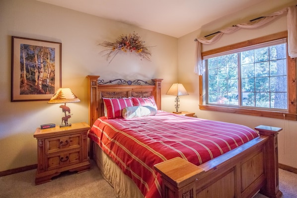 A bedroom with a wooden bed, two bedside tables, a nightstand, a window with curtains, and a wall-mounted artwork.
