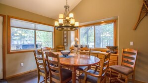 A dining area with a round wooden table, six chairs, a chandelier, and large windows offering a view of trees. - Red Cedar 49 (Bend)