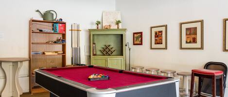 A pool table with a red felt surface, surrounded by stools and a green cabinet with decorative items.