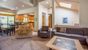 A spacious living room with a large brown leather sofa, a smaller armchair, and a coffee table. There is a dining area with a wooden table and chairs, and a kitchen in the background. - Sumac 9 (Bend)