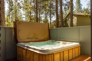 A hot tub with a wooden exterior, surrounded by a wooden deck and a wooden fence, with a house and trees in the background. - Tan Oak 29 (Bend)