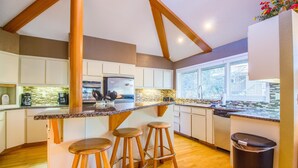 A modern kitchen with wooden beams, a central island with stools, and a view of the outdoors through large windows. - Tan Oak 15 (Bend)