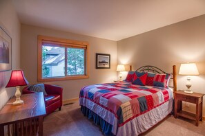 Quarto com uma cama grande, duas mesas de cabeceira com luminárias, uma cadeira vermelha, uma janela com vista para uma casa e um quadro na parede.