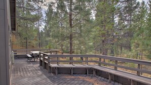 Una terraza de madera con bancos y mesas, rodeada de altos pinos.