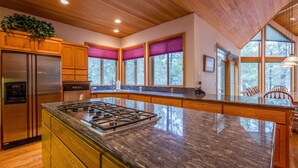 A modern kitchen with wooden cabinets, a stainless steel refrigerator, a granite countertop, and large windows with purple blinds.