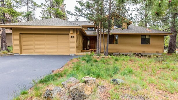 Une maison à deux étages avec un garage, entourée de pins et d’une zone herbeuse parsemée de rochers.