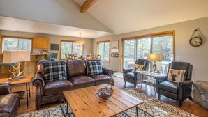 A spacious living room with a brown leather sofa, a wooden coffee table, and a stone fireplace. There are two armchairs and a dining table with chairs. The room has large windows and a chandelier. - Topflite 12 (Bend)