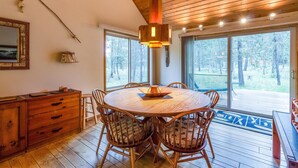 Uma mesa de jantar de madeira com cadeiras, um lustre pendente e vista para o exterior através de grandes janelas.