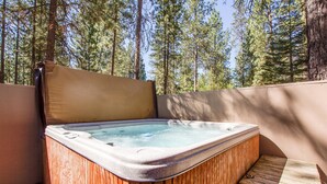 A hot tub with a wooden exterior and a cushioned seat, surrounded by a wooden deck and a wooden fence, with a forest in the background. - Tumalo 11 (Bend)