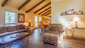 A cozy living room with a wooden ceiling, a fireplace, a sofa, a patterned armchair, and a dining area with wooden furniture. - Tumalo 11 (Bend)