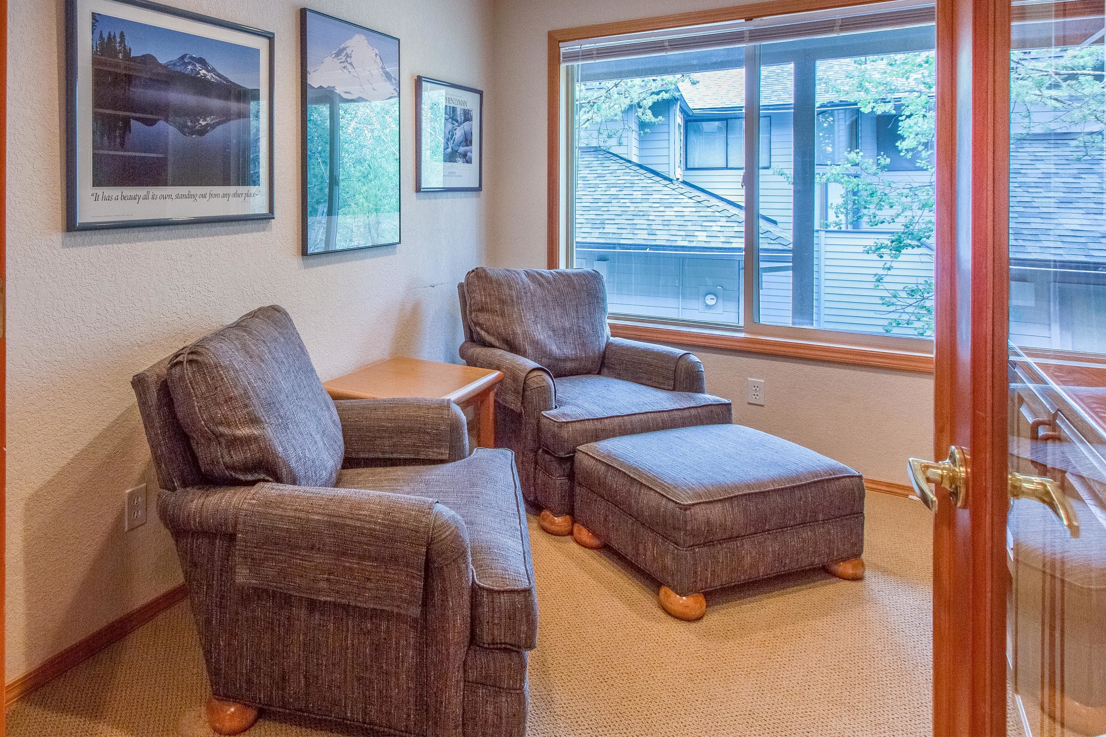 A room with two armchairs and a small table, a large window with a view of a house, and framed pictures on the wall.