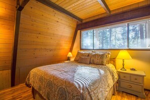 Un dormitorio acogedor con techo de madera, una cama grande con ropa de cama estampada, una mesita de noche con lámpara y una ventana con persianas.