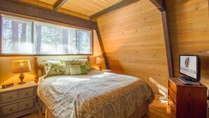 A cozy bedroom with a wooden ceiling, a large bed with patterned bedding, a nightstand with a lamp, and a television on a wooden stand. - Woodland 1 (Bend)