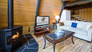 A cozy room with a fireplace, a television on a wooden stand, a white sofa with blue cushions, and a staircase leading to an upper level. - Woodland 1 (Bend)
