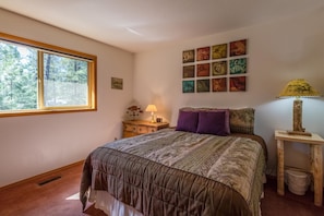 A bedroom with a large window, a bed with a patterned bedspread, a nightstand with a lamp, and a wall with framed artwork. - Woodland 7 (Bend)