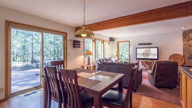 Un comedor con mobiliario de madera, un ventanal con vistas a árboles y un televisor mostrando 'BENNINGTON PROPERTIES'.