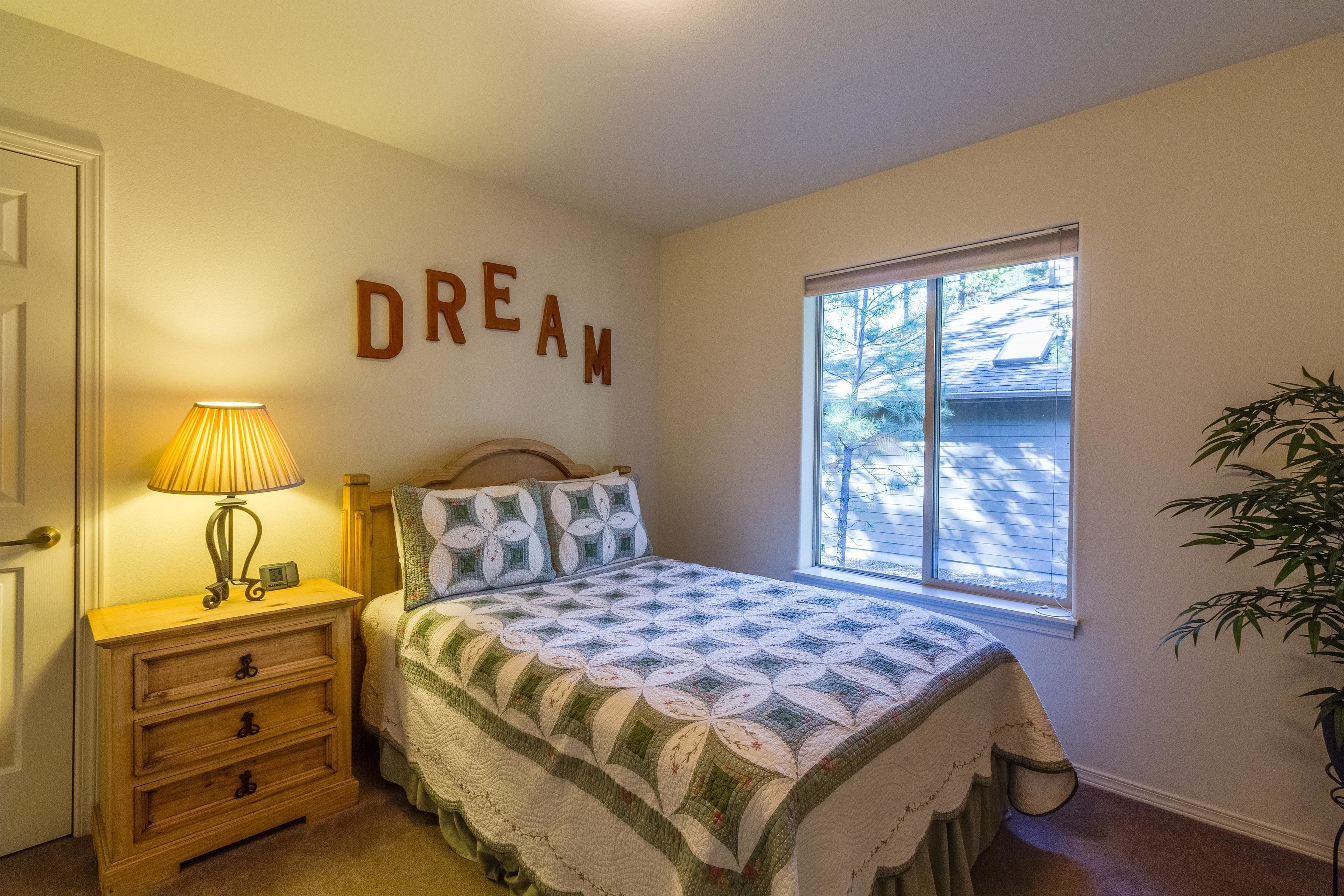 A bedroom with a bed, a nightstand with a lamp, a wall with the word "DREAM" spelled out, and a window with a view of trees.