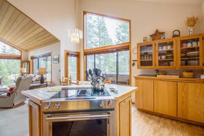 A modern kitchen with a central island, wooden cabinets, and large windows offering views of trees.