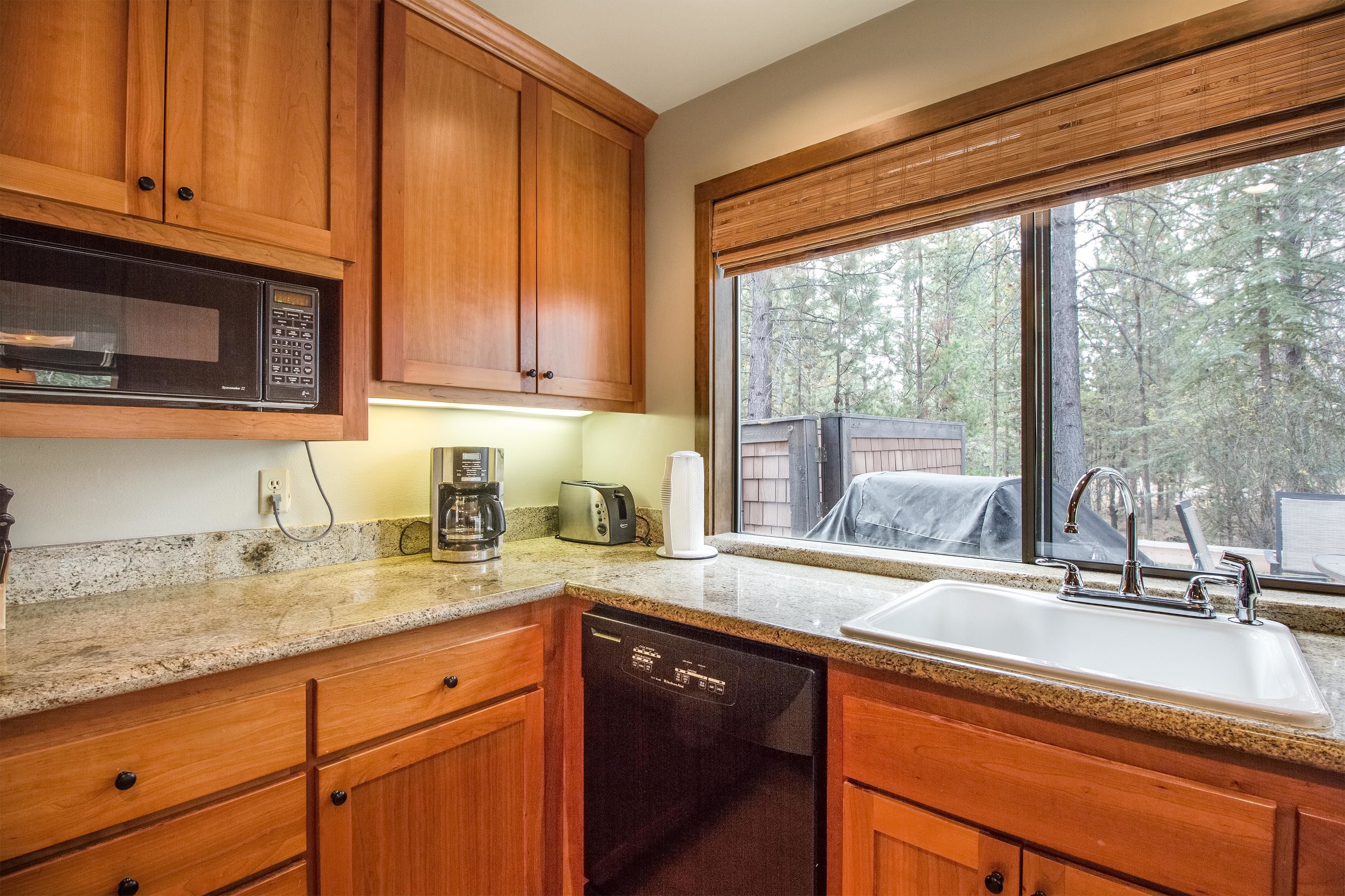 A kitchen with wooden cabinets, a built-in microwave, a dishwasher, a sink, and a window with a view of trees.