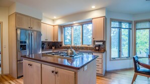 A modern kitchen with wooden cabinets, stainless steel appliances, a central island, and large windows offering natural light and views. - Siskin 12 (Bend)