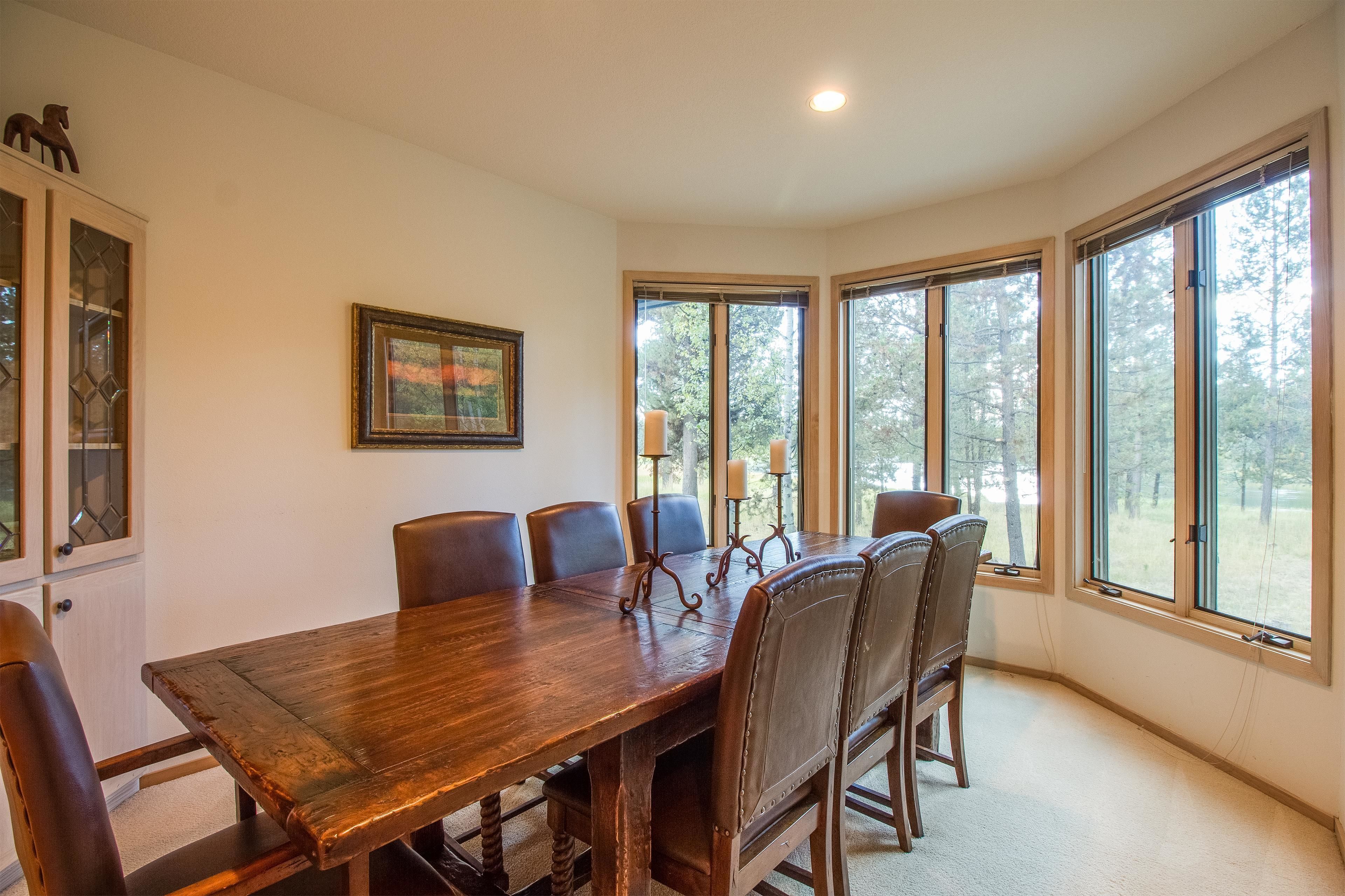 A dining room with a wooden table and chairs, large windows with a view of trees, a cabinet with glass doors, and a framed picture on the wall.