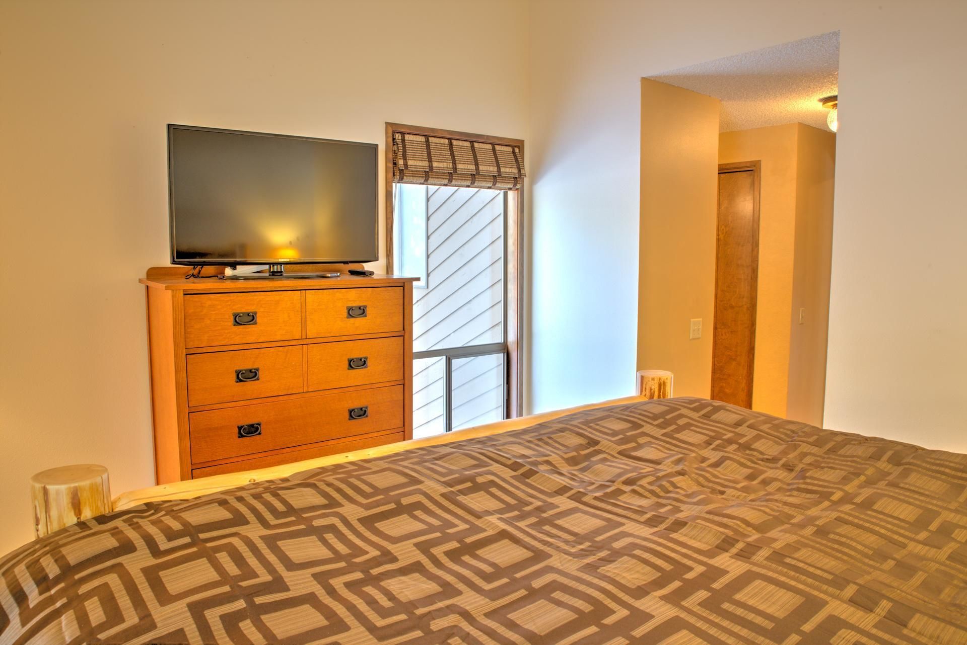 A bedroom with a bed, a wooden dresser with a television on top, a window with blinds, and a door.