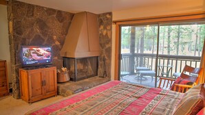 A bedroom with a stone fireplace, a television, a large bed with a patterned blanket, and a balcony with outdoor furniture.