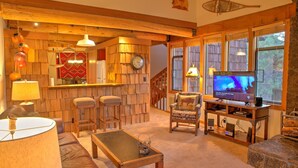 A cozy living room with a wooden bar, comfortable seating, a television, and large windows.