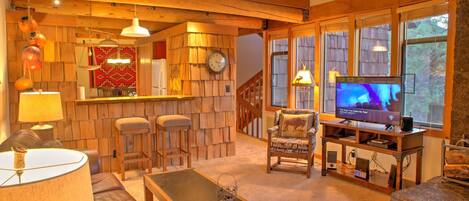 A cozy living room with a wooden bar, comfortable seating, a television, and large windows.