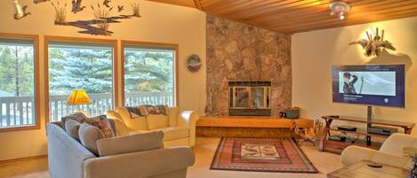 A living room with a stone fireplace, a television, and a large window overlooking a forest.
