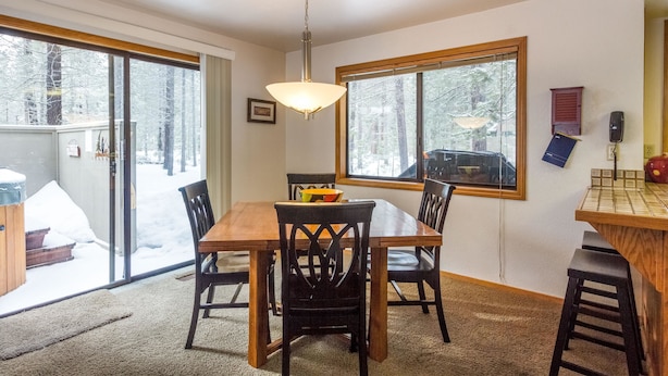 A dining area with a wooden table and chairs, a hanging light fixture, and large windows with a view of a snowy landscape. - Backwoods 3 (Bend)