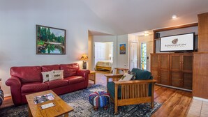 A living room with a red leather sofa, a wooden coffee table, and a blue ottoman. There is a television on a wooden cabinet and a painting on the wall. - Kinglet 23 (Bend)