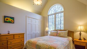 A bedroom with a large window, a wooden dresser, a bed with floral bedding, and two bedside lamps. - Kinglet 23 (Bend)