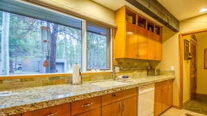 A kitchen with granite countertops, wooden cabinets, a built-in dishwasher, and a large window overlooking a wooded area. - Meadow House Cndo 8 (Bend)