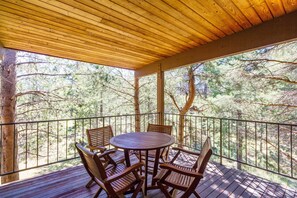 Una terraza de madera con una mesa redonda y cuatro sillas, rodeada por una barandilla de metal, con vistas a un área boscosa.