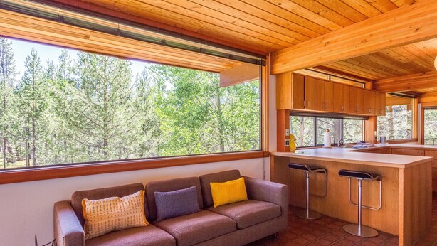 A modern kitchen with a wooden ceiling, a large window overlooking a forest, a bar area with stools, and a sofa with cushions. - Wickiup 14 (Bend)