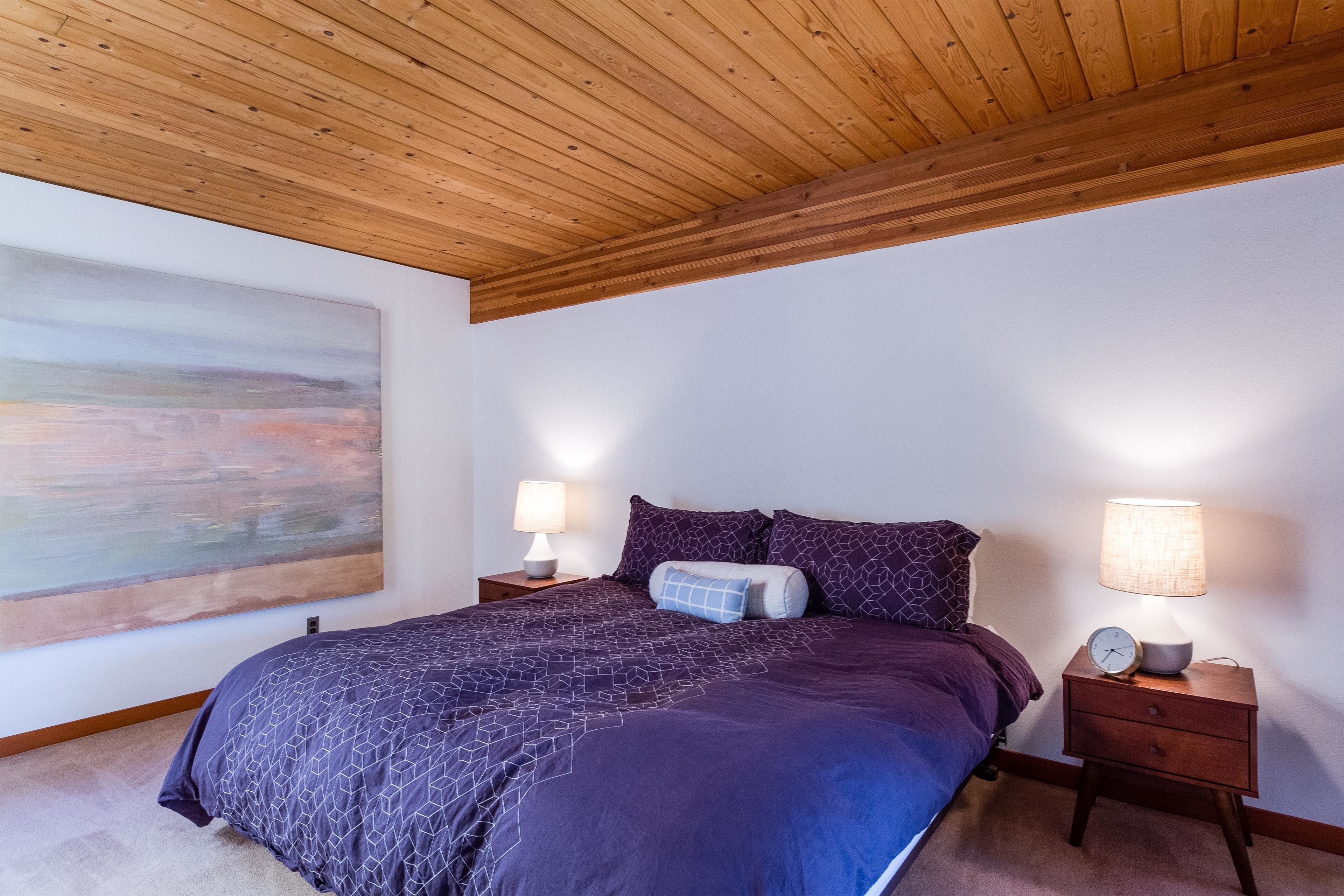 A bedroom with a wooden ceiling, a large abstract painting, a bed with a purple geometric bedspread, two bedside tables with lamps, and a clock.