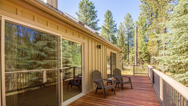 Une terrasse en bois avec des chaises et une porte vitrée donnant sur un espace clos, entouré d’arbres.