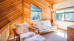 Une pièce chaleureuse aux murs en bois et au plafond en pente, comprenant un canapé, un fauteuil, une lampe et une fenêtre donnant sur les arbres.