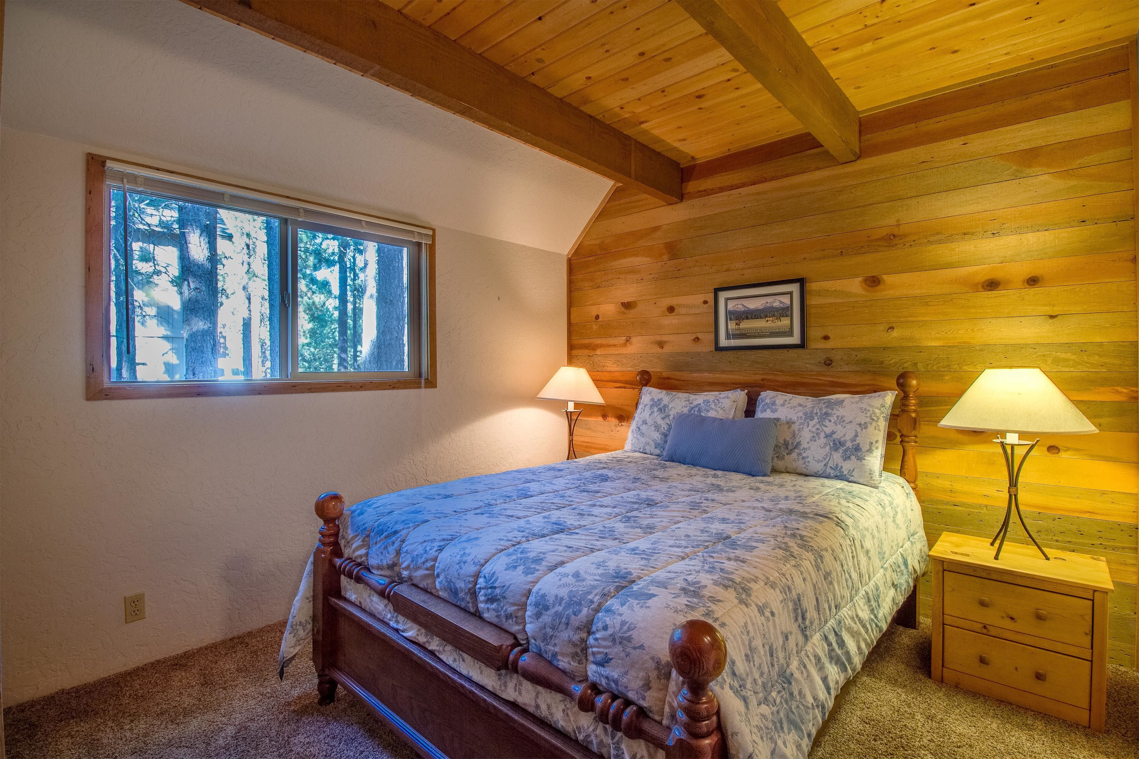 Ein Schlafzimmer mit Holzboden, einem großen Fenster, einem Bett mit blumigem Überwurf, zwei Nachttischen mit Lampen und einem gerahmten Bild an der Wand.