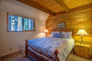 A bedroom with a wooden ceiling, a large window, a bed with a floral bedspread, two bedside tables with lamps, and a framed picture on the wall. - Mink 3 (Bend)