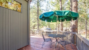 A wooden deck with a table and chairs under a green umbrella, surrounded by trees. - Ashwood 4 (Bend)
