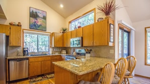 A modern kitchen with wooden cabinets, granite countertops, a large island with bar stools, and a view of the outdoors through large windows. - Ashwood 4 (Bend)